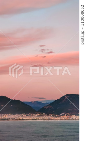 Pink clouds in evening sky over blue sea on background of city and mountains on horizon, vertical frame. Pink clouds in evening sky over blue sea on background of city and mountains on horizon, vertical frame. 110008052