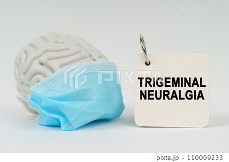 On a white surface there is a brain with a blue mask and a notepad with the inscription - Trigeminal neuralgia On a white surface there is a brain with a blue mask and a notepad with the inscription - Trigeminal neuralgia 110009233