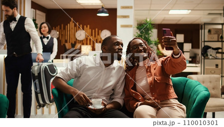 Newlywed couple in video conference with relatives while on honeymoon trip. Smiling husband in video call with parents in law, enjoying cup of coffee in hotel lounge, zoom out shot 110010301