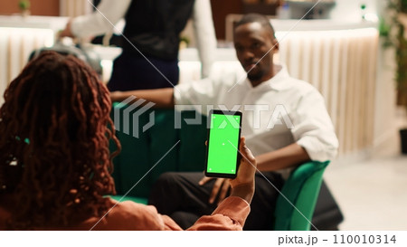 Company partners away on business trip waiting in hotel foyer to be checked in, holding chroma key green screen mock up smartphone. Coworkers chatting in cozy resort lobby, checking info on phone 110010314