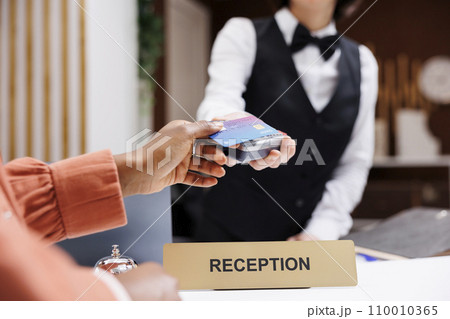 Female tourist paying at pos terminal in hotel lobby, using electronic payment at check in registration. Young resort guest using credit card at reception counter front desk. Close up. 110010365