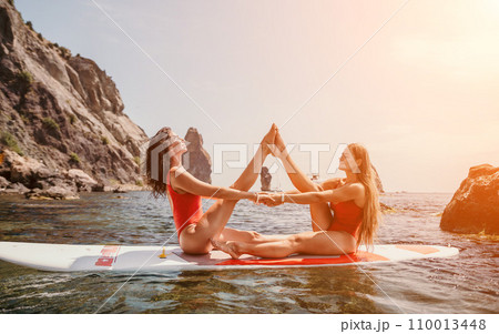 Woman sup yoga. Two happy sporty women practising yoga pilates on paddle sup surfboard. Female stretching doing workout on sea water. Modern individual female hipster outdoor summer sport activity. 110013448
