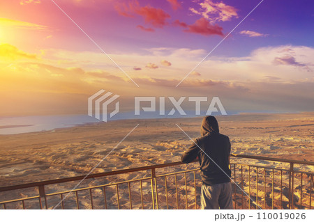Young man standing on a viewpoint on the mount (Masada) and gazing sunrise over the Dead Sea. Dead sea region, Masada, Israel 110019026