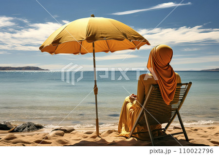 Muslim woman with hijab sits next to a beach umbrella on a sun lounger at a tranquil coast 110022796