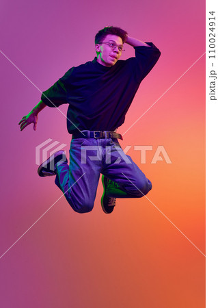 Young man in casual stylish clothes and eyeglasses jumping with positive face against gradient pink background in neon light 110024914