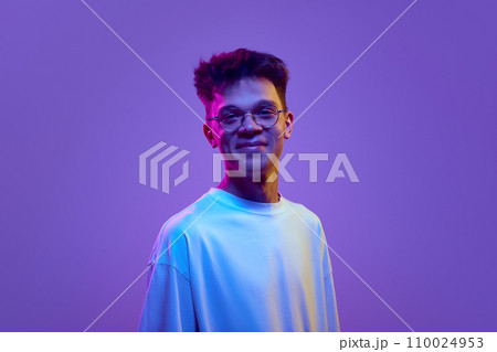 Portrait of smiling young man, boy in casual white t-shirt and sunglasses looking at camera against purple background in neon light Portrait of smiling young man, boy in casual white t-shirt and sunglasses looking at camera against purple background in neon light 110024953