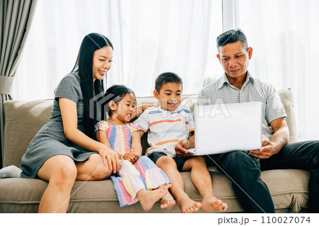 Parents and children happily share a laptop on a sofa browsing contentedly. This image portrays familial bonding joy and the wonder of shared technology experiences within the family. 110027074