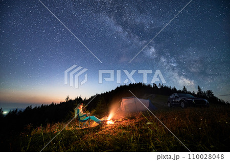 Night camping in the mountains under starry sky full of stars. Travelling woman admiring starry night in mountains. Young woman sitting next to camp fire, tourist tent and off-road car. Night camping in the mountains under starry sky full of stars. Travelling woman admiring starry night in mountains. Young woman sitting next to camp fire, tourist tent and off-road car. 110028048