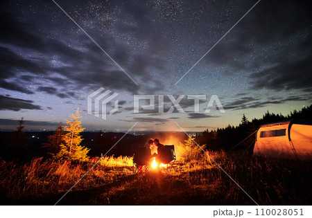 Romantic couple enjoying at starry sky. Young people sitting in chairs and warming by campfire near tourist tent. Travelling couple having a rest together. Night camping. Romantic couple enjoying at starry sky. Young people sitting in chairs and warming by campfire near tourist tent. Travelling couple having a rest together. Night camping. 110028051