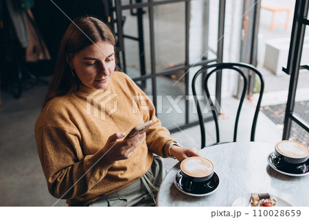 Beautiful young woman in casual clothes smiling, drinking a cup of coffee and holding a phone in her hands while spending time in the cafe. Modern interior. Food concept 110028659