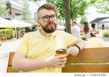 Millennial smiling student man in yellow polo t-shirt eyeglasses sitting on bench drink coffee have breakfast rest relax in sunshine spring green city park outdoors on nature. Urban leisure concept. Millennial smiling student man in yellow polo t-shirt eyeglasses sitting on bench drink coffee have breakfast rest relax in sunshine spring green city park outdoors on nature. Urban leisure concept. 110029186
