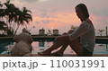 Woman relaxes sitting near pool on tropical island hotel resort. Bright colorful sunset sky and coconut palms reflected in water. Travel, tourism, holiday spa vacation. 110031991