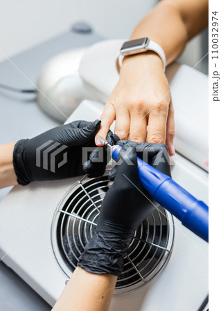 Manicurist uses an electric drill to remove old gel polish from nails. A woman is getting a manicure of nails. The beautician files the client's nails. professional manicure tool. Close-up 110032974
