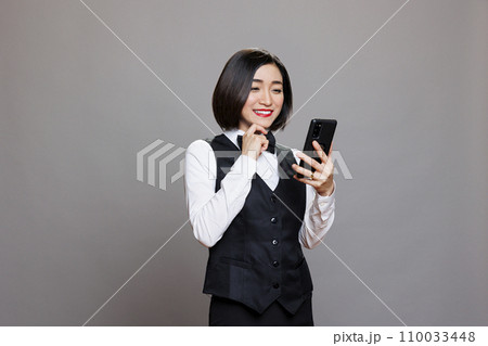 Happy asian cafe waitress reading client positive feedback on smartphone. Cheerful smiling receptionist in professional uniform checking customer satisfaction while using mobile phone Happy asian cafe waitress reading client positive feedback on smartphone. Cheerful smiling receptionist in professional uniform checking customer satisfaction while using mobile phone 110033448