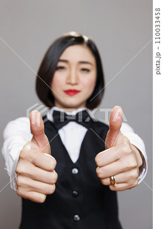 Cheerful asian woman in waitress uniform smiling and giving thumbs up symbol closeup. Cafe worker confirming and recommending restaurant good service and looking at camera Cheerful asian woman in waitress uniform smiling and giving thumbs up symbol closeup. Cafe worker confirming and recommending restaurant good service and looking at camera 110033458