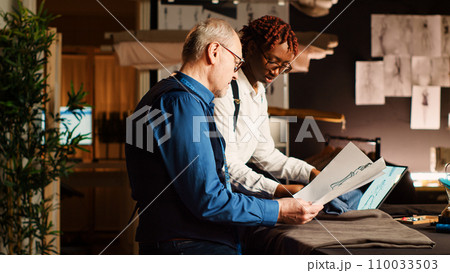 Experienced tailor and student working in atelier, looking at clothes design sketches to craft fashion collection. Apprentice learning the art of sewing from senior couturier. Handheld shot. Experienced tailor and student working in atelier, looking at clothes design sketches to craft fashion collection. Apprentice learning the art of sewing from senior couturier. Handheld shot. 110033503