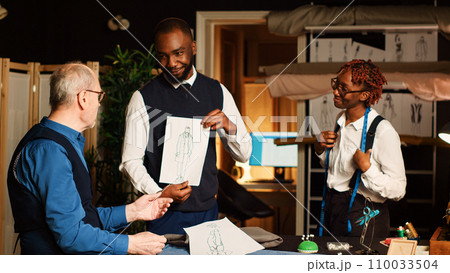 Customer discussing about sizes with senior couturier and assistant to design suit, woman taking measurements with meter. Measuring tape and manufacturing tools in atelier workshop. Customer discussing about sizes with senior couturier and assistant to design suit, woman taking measurements with meter. Measuring tape and manufacturing tools in atelier workshop. 110033504