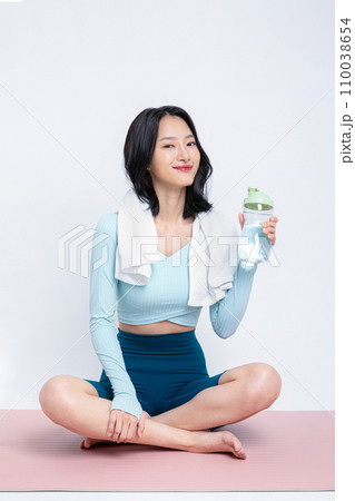 There is a woman sitting on a yoga mat with a water bottle. There is a woman sitting on a yoga mat with a water bottle. 110038654