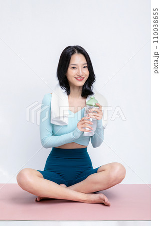 There is a woman sitting with a towel on a yoga mat with a water bottle in her hand 110038655