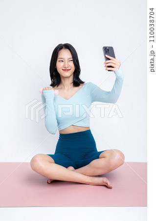 There is a woman sitting on a yoga mat and taking selfies with her phone. 110038704