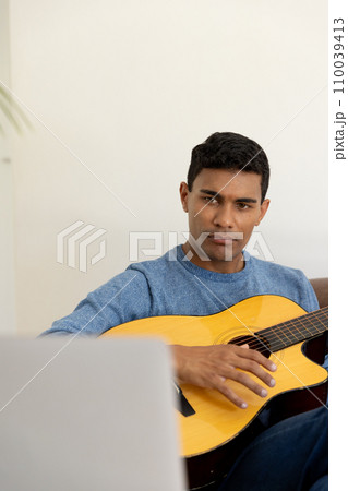 Biracial man sitting on sofa playing guitar and using laptop at home 110039413