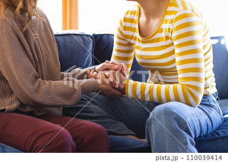 Midsection of biracial lesbian couple holding hands sitting on couch at home 110039414