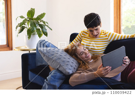 Happy biracial lesbian couple relaxing on couch together using tablet at home, copy space 110039422