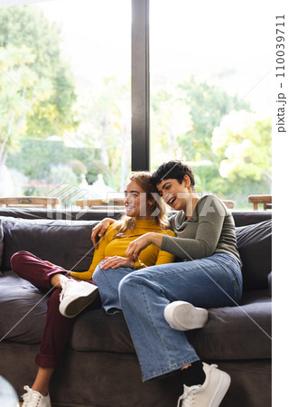 Happy biracial lesbian couple sitting on couch laughing in sunny living room, copy space Happy biracial lesbian couple sitting on couch laughing in sunny living room, copy space 110039711