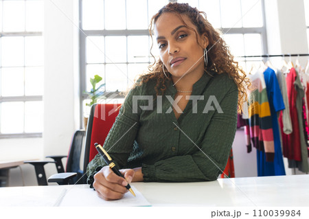 Focused biracial female fashion designer having video call and taking notes at desk in studio Focused biracial female fashion designer having video call and taking notes at desk in studio 110039984