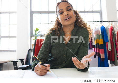 Happy biracial female fashion designer having video call and taking notes at desk in studio 110039998