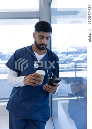Biracial male doctor using smartphone and holding cup of coffee in hospital office Biracial male doctor using smartphone and holding cup of coffee in hospital office 110040430
