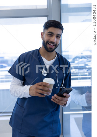 Portrait of happy biracial male doctor using smartphone and holding cup of coffee in hospital office 110040431