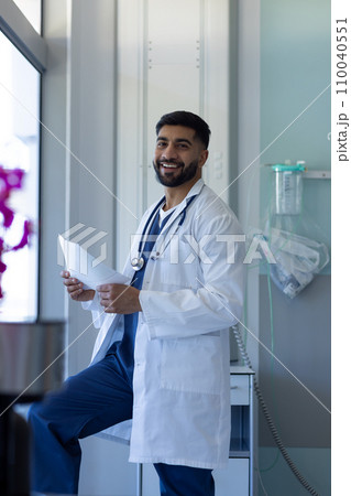 Portrait of happy biracial male doctor reading document in sunny hospital room Portrait of happy biracial male doctor reading document in sunny hospital room 110040551