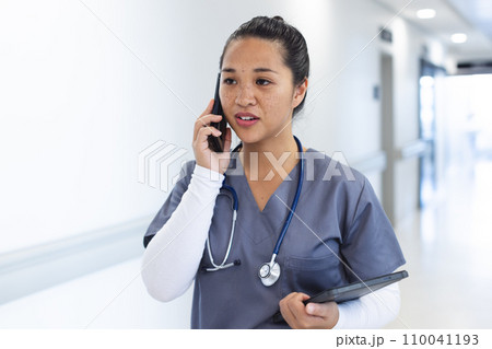 Biracial female doctor wearing scrubs talking on smartphone in hospital corridor 110041193