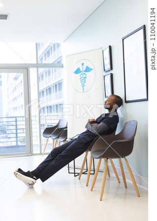 African american male doctor wearing scrubs sitting in waiting room in hospital, copy space 110041194
