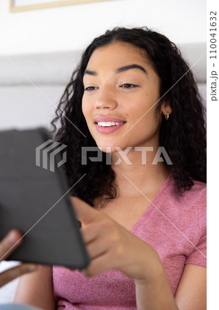 Happy biracial woman using tablet sitting on bed at sunny home 110041632