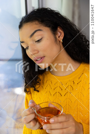 Thoughtful biracial woman holding cup of tea and looking out window at home Thoughtful biracial woman holding cup of tea and looking out window at home 110041647