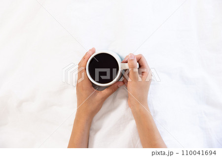 Hands of biracial woman holding cup of coffee sitting in bed at sunny home Hands of biracial woman holding cup of coffee sitting in bed at sunny home 110041694
