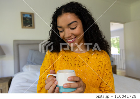 Happy biracial woman in yellow sweater holding cup of coffee at home 110041729