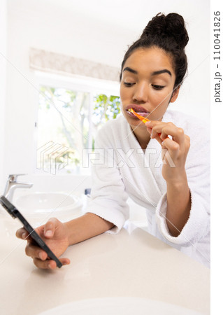Biracial woman in bathrobe brushing teeth and using smartphone in sunny bathroom 110041826