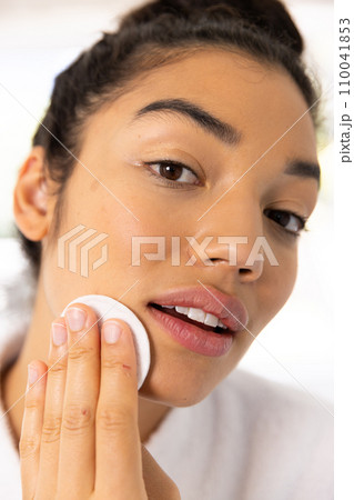 Portrait of biracial woman in bathrobe cleansing her face in sunny bathroom Portrait of biracial woman in bathrobe cleansing her face in sunny bathroom 110041853