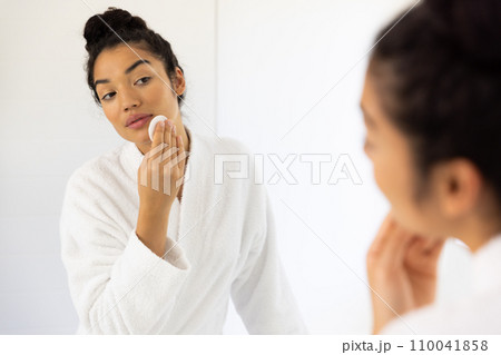 Biracial woman in bathrobe cleansing her face in sunny bathroom 110041858