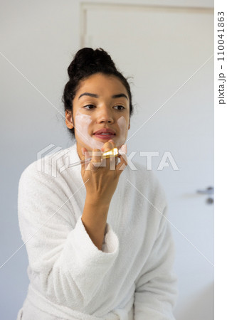 Biracial woman in bathrobe applying face mask using brush in sunny bathroom Biracial woman in bathrobe applying face mask using brush in sunny bathroom 110041863