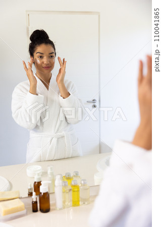 Biracial woman in bathrobe applying cream on her face in sunny bathroom Biracial woman in bathrobe applying cream on her face in sunny bathroom 110041865