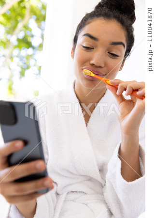 Biracial woman in bathrobe brushing teeth and using smartphone in sunny bathroom Biracial woman in bathrobe brushing teeth and using smartphone in sunny bathroom 110041870