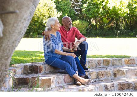 Happy diverse senior couple embracing and sitting on stairs in sunny garden 110042430