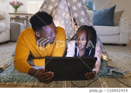 Happy african american father and daughter lying by teepee with tablet at home, copy space 110042454