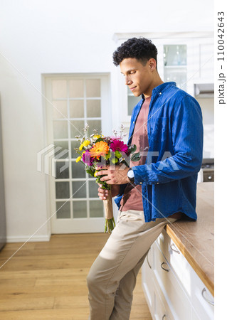 Happy biracial man holding flowers in kitchen at home, copy space 110042643