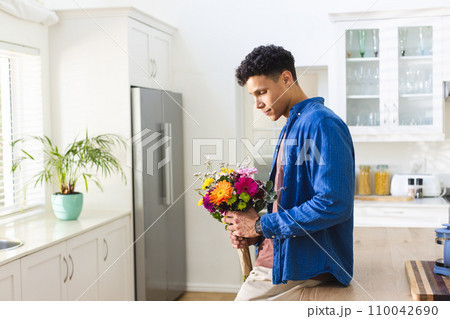 Happy biracial man holding flowers in kitchen at home, copy space 110042690
