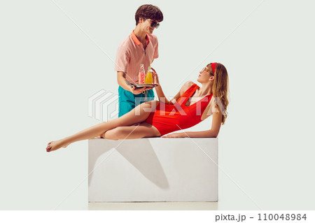 Summer seaside resort. Young man serving drink to beautiful woman in swimsuit sunbathing against white background 110048984